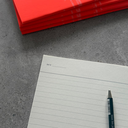 Everyday Notebook open on a lined page with a DATE field, a pen, and a stack of orange notebooks behind