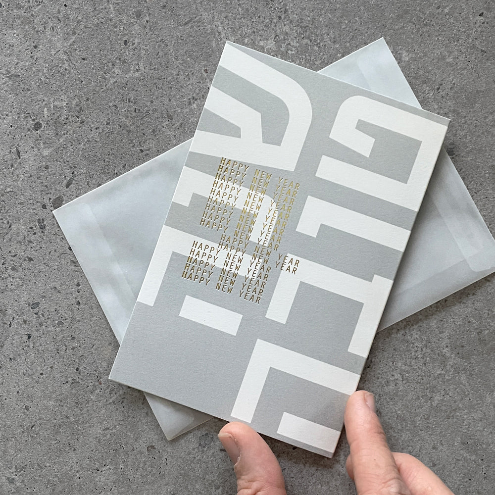 Grey Shana Tova greeting card with gold Happy New Year lettering, held above its translucent envelope