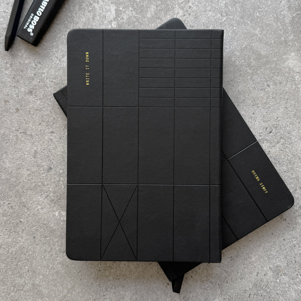 Black PU reversible hardcover notebook with embossed geometric line pattern, Write It Down gold foil stamp on top notebook and Doing Stuff stamp on flipped notebook below