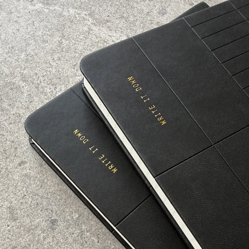 Two black PU notebooks stacked showing Write It Down foil stamp on both covers
